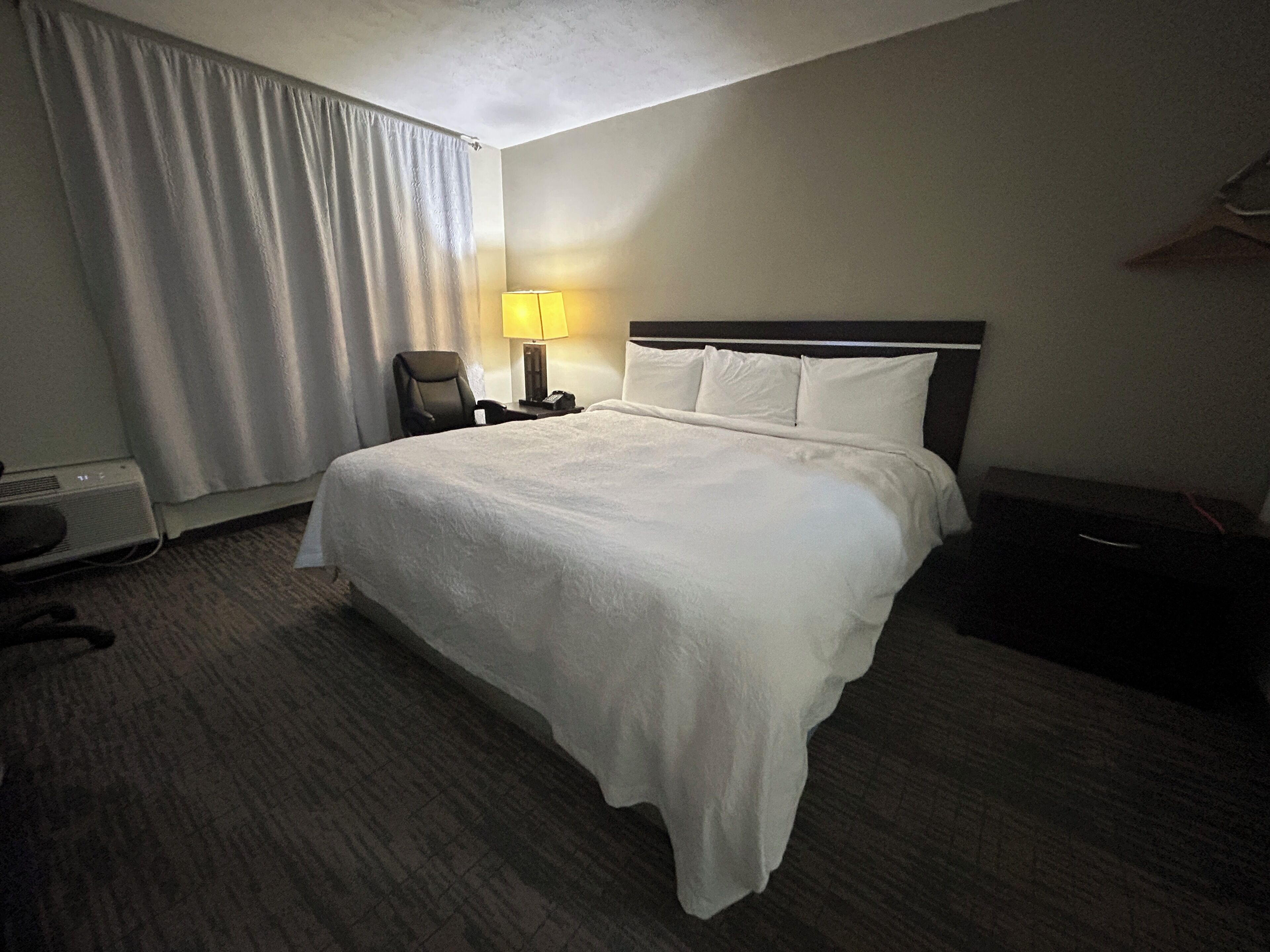 A hotel room with a large bed, a desk with a lamp, a chair, and a coat rack.