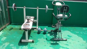 A weight bench with a barbell and weights, a squat rack, and an elliptical machine in a room with green walls and a green floor. - Gumi A Motel (Gumi)