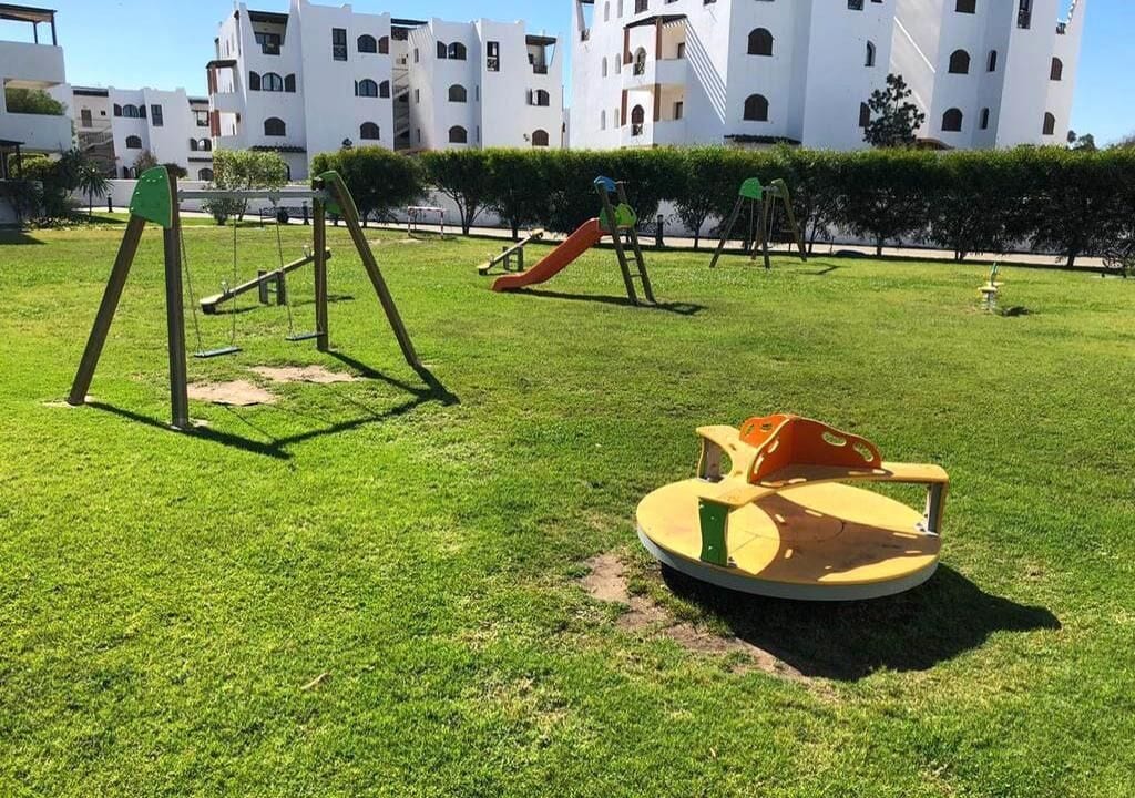 Un terrain de jeux avec des balançoires et un toboggan, entouré de pelouse bien entretenue et avec un bâtiment en arrière-plan.