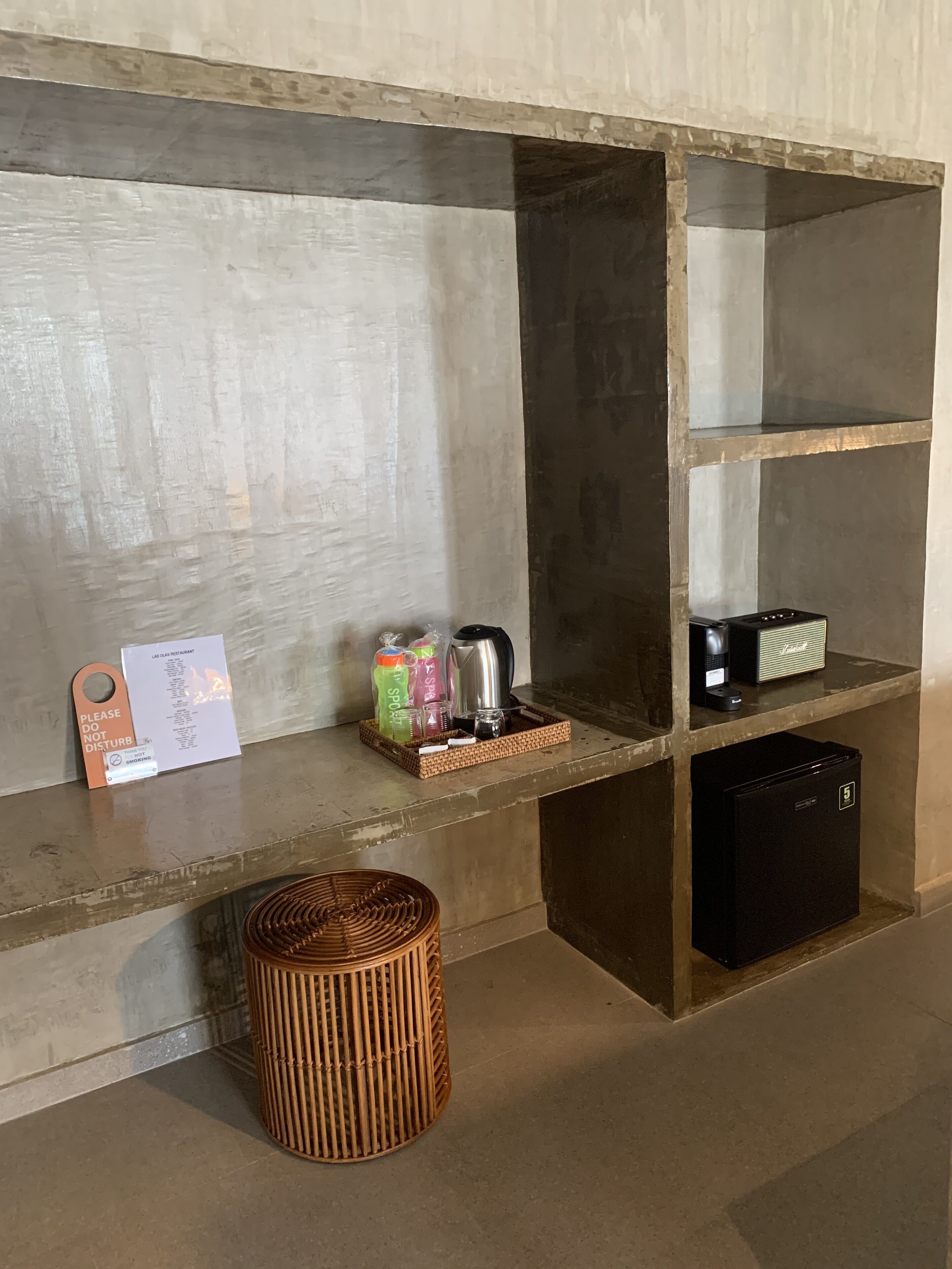 Habitación de hotel moderna y limpia, con una pared de concreto decorativo, una pequeña repisa con un cartel, una cesta y un refrigerador negro.