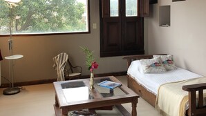 Une pièce chaleureuse avec un plafond en bois, une grande fenêtre, un lit agrémenté d’oreillers à motifs floraux, une petite table sur laquelle sont posés un vase et des magazines, et un lampadaire.