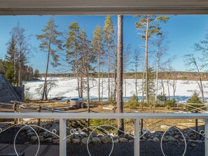 Eine Holzterrasse mit Geländer, mit Blick auf eine verschneite Landschaft mit Bäumen und einem Gewässer.