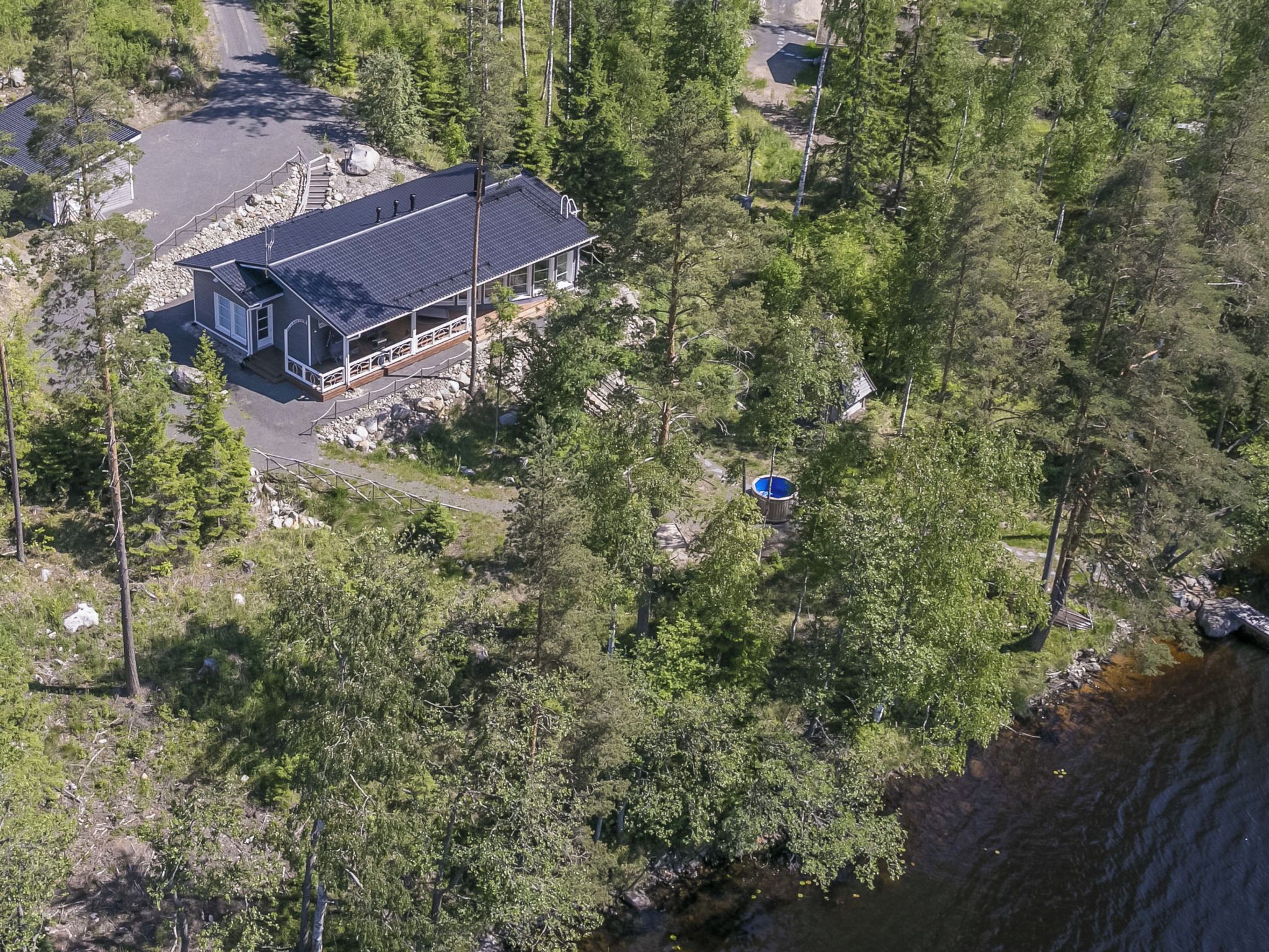 Kaksikerroksinen talo, jossa on parveke, ja sen ympärillä on tiheää metsää sekä vesistö.