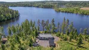 En stuga vid sjön med ett tak av metall, omgiven av tät skog och en stor sjö.