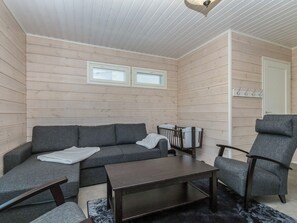 Ein Zimmer mit einer Couch, einem Sessel, einem Tisch und einem Babybett. Zwei Fenster mit Jalousien sind vorhanden.