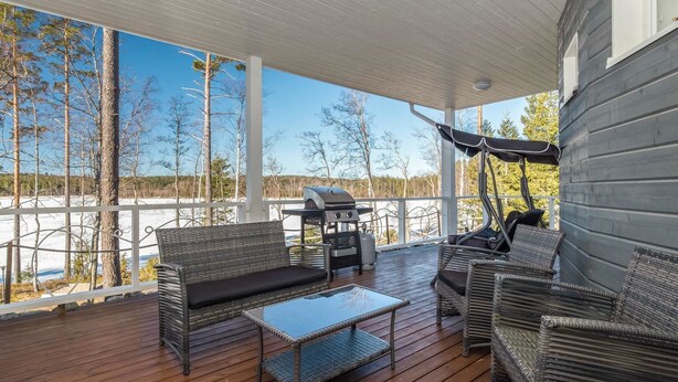 En takförsedd uteplats med möbler i rotting, en grill och utsikt över ett snötäckt landskap.
