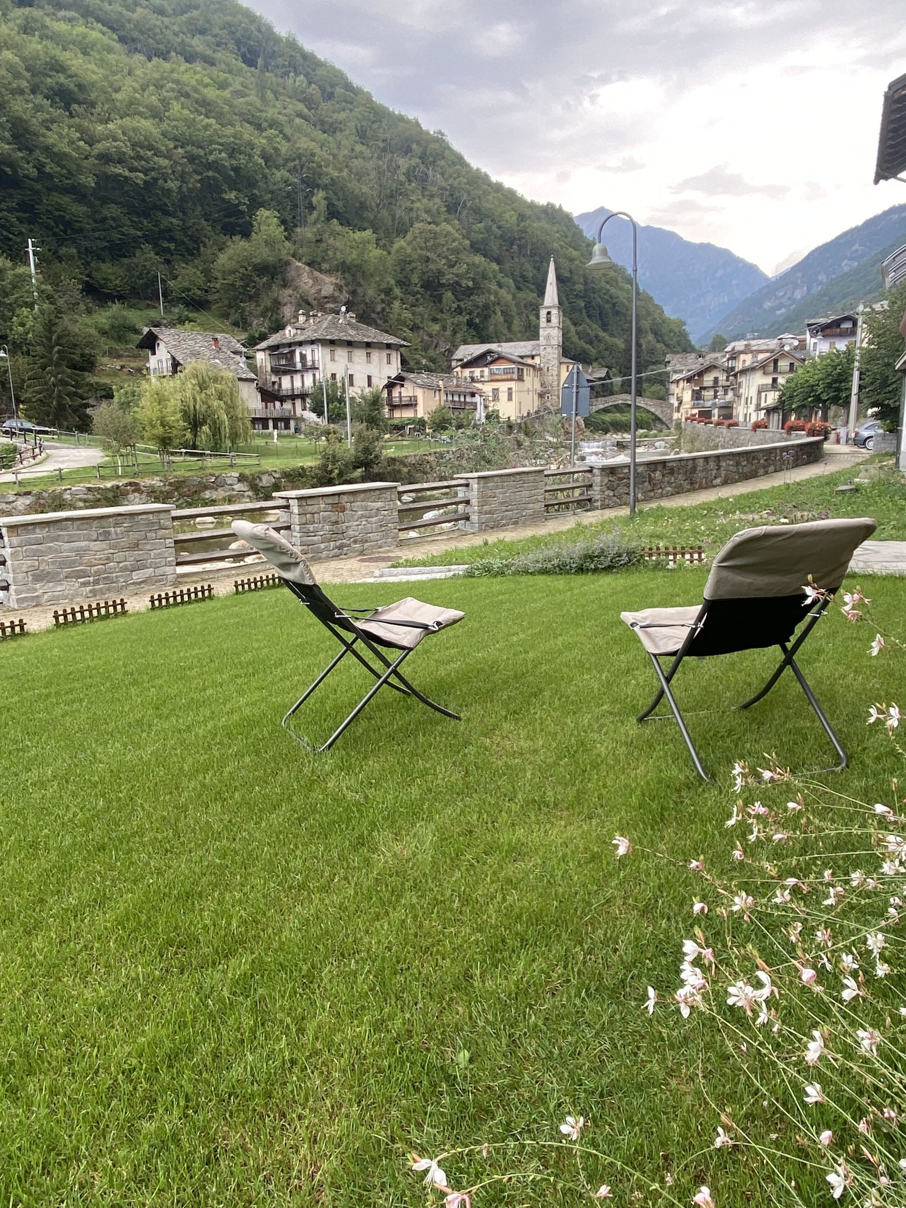 A grassy outdoor area with two folding chairs, a stone wall, and a view of a village with buildings and a church.