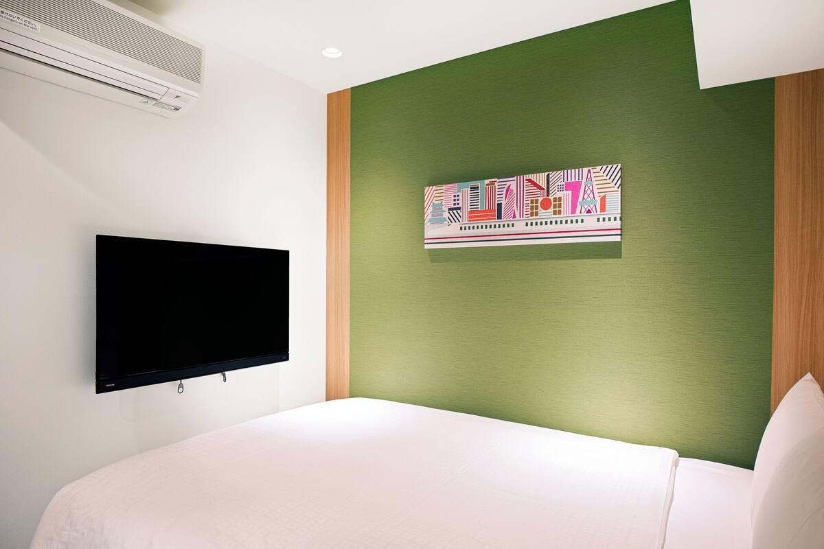 A hotel room with a bed, a flat-screen TV, an air conditioner, and a green wall with a colorful abstract artwork.