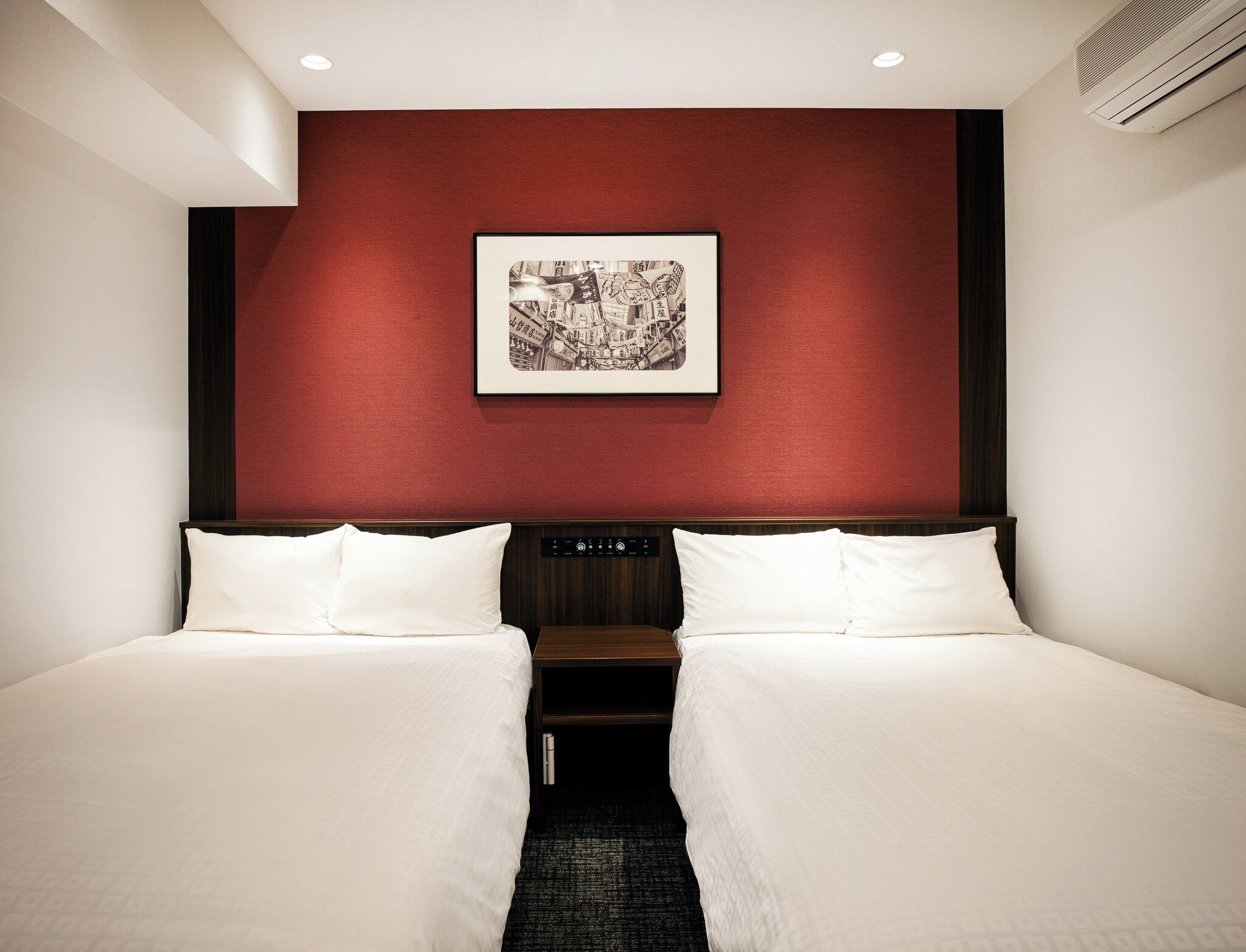 Habitación de hotel con dos camas, una pared de acento roja, un cuadro enmarcado y un panel de control.