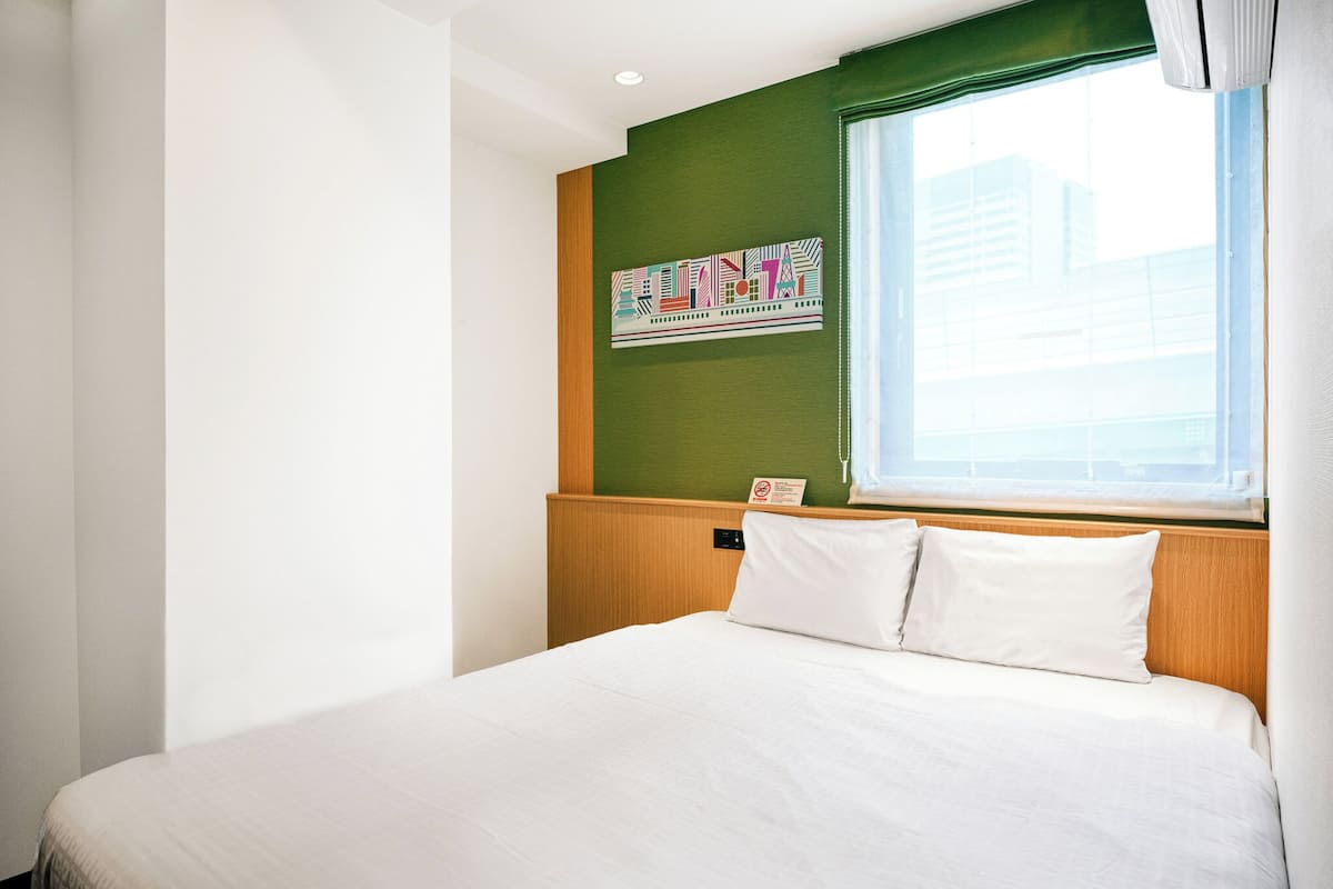 A hotel room with a bed, a green accent wall, a window with blinds, and a wall-mounted artwork.