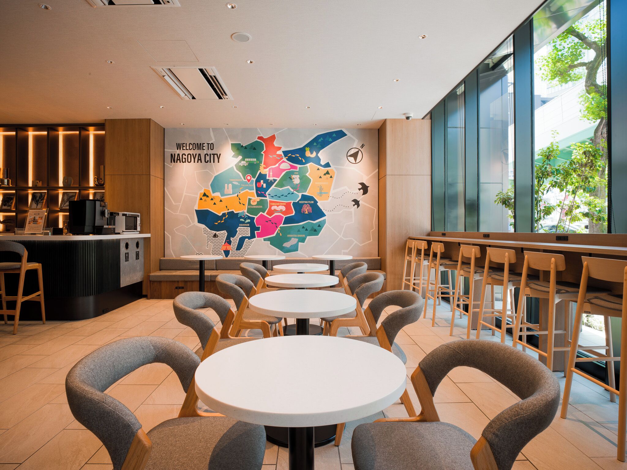 Ein modernes Café-Interieur mit runden Tischen, grauen Stühlen, einer Wand mit einer Karte und Blick ins Grüne.
