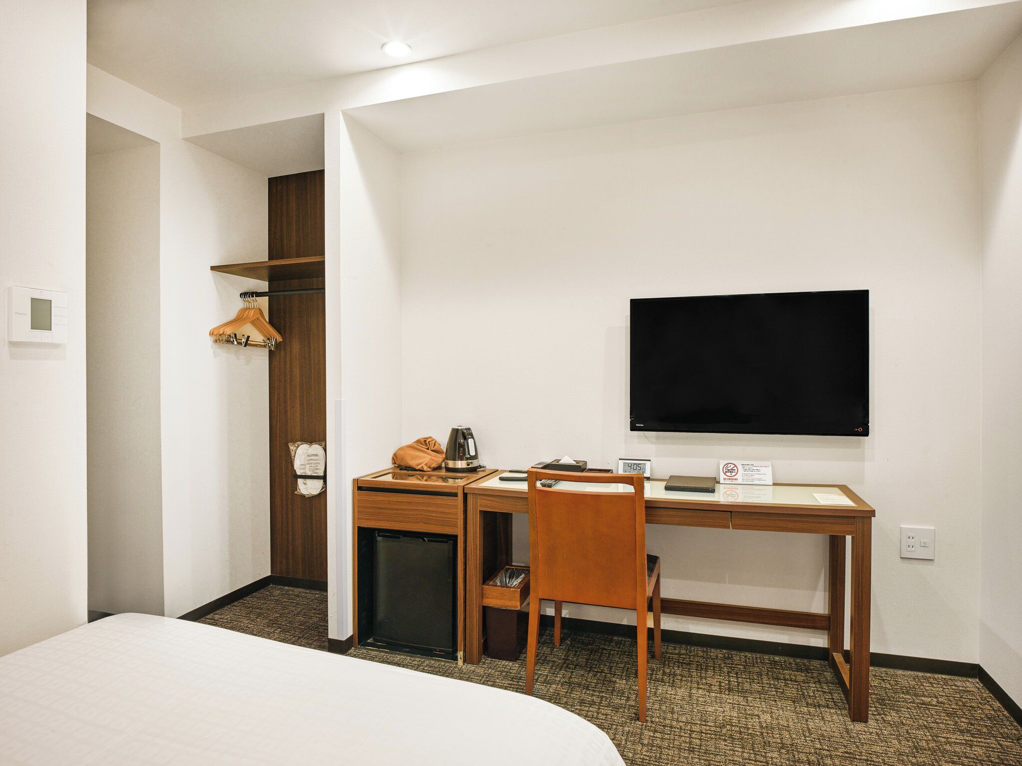 Habitación de hotel con cama, escritorio con silla, televisor, perchero y refrigerador pequeño.