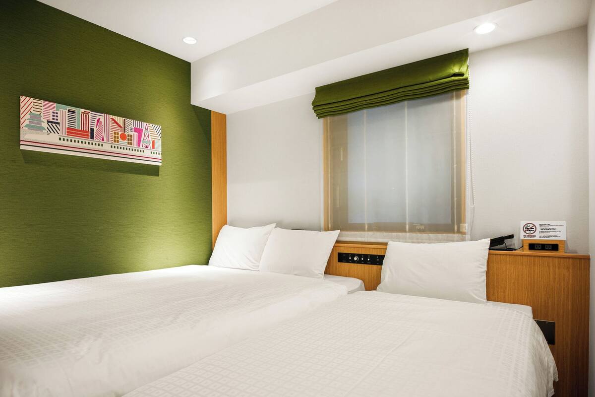 A hotel room with two beds, a green accent wall, a window with a green valance, and a bedside table with a clock.