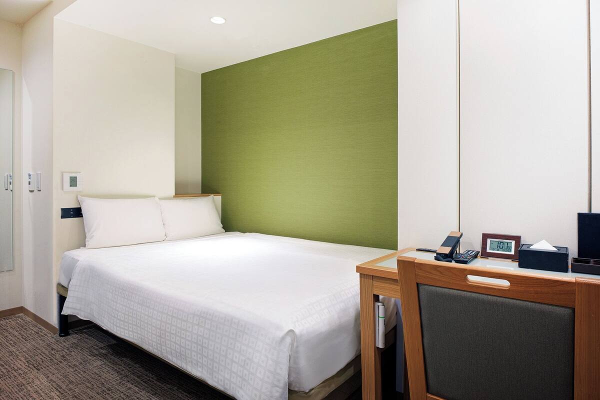 A hotel room with a bed, a desk with a telephone, a digital clock, and a thermostat on the wall.