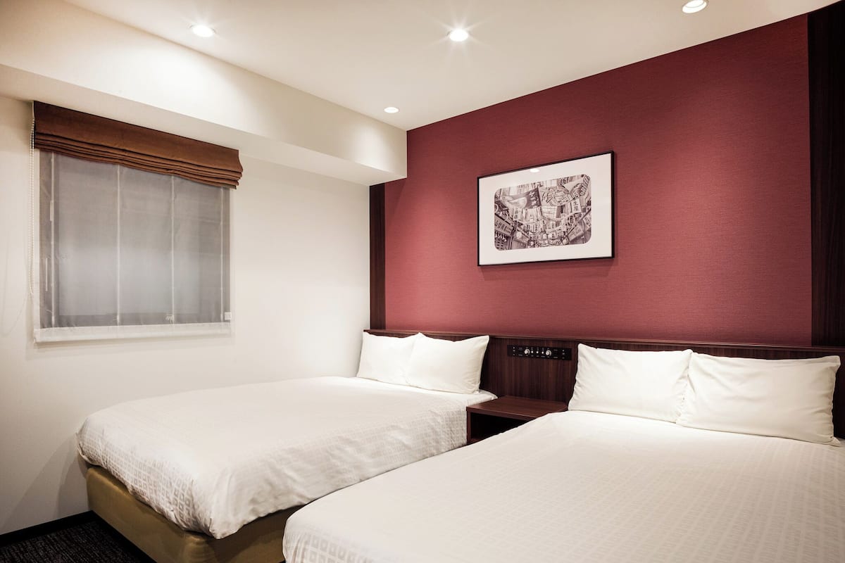 A hotel room with two beds, a window with blinds, a wall-mounted control panel, and a framed picture.