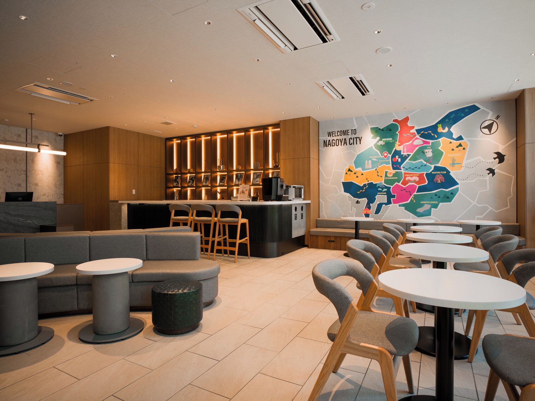 A modern hotel lobby with a reception desk, seating area, and a map of Nagoya City on the wall.