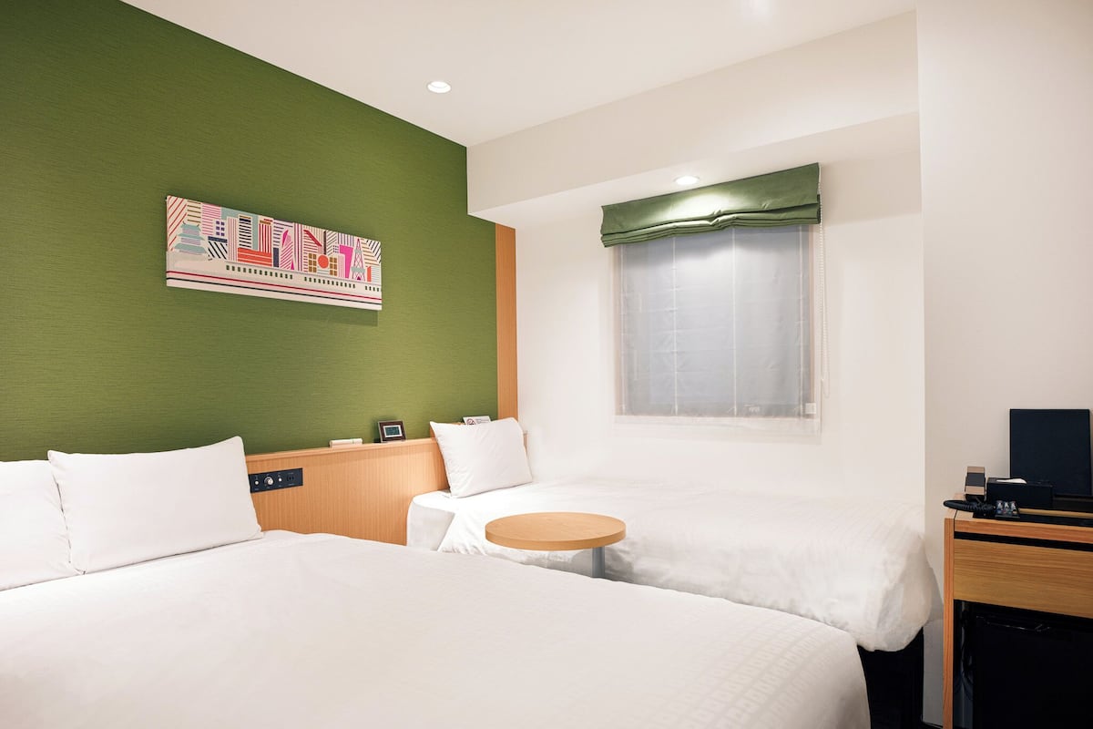 A hotel room with two beds, a small round table, a green accent wall, and a window with a green valance.