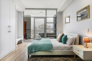 A modern bedroom with a large window, a bed with multiple pillows, a bedside table with a lamp, and a view of the cityscape.