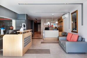 A modern hotel lobby with a reception desk, a seating area with a sofa and armchairs, and a kitchen area in the background.