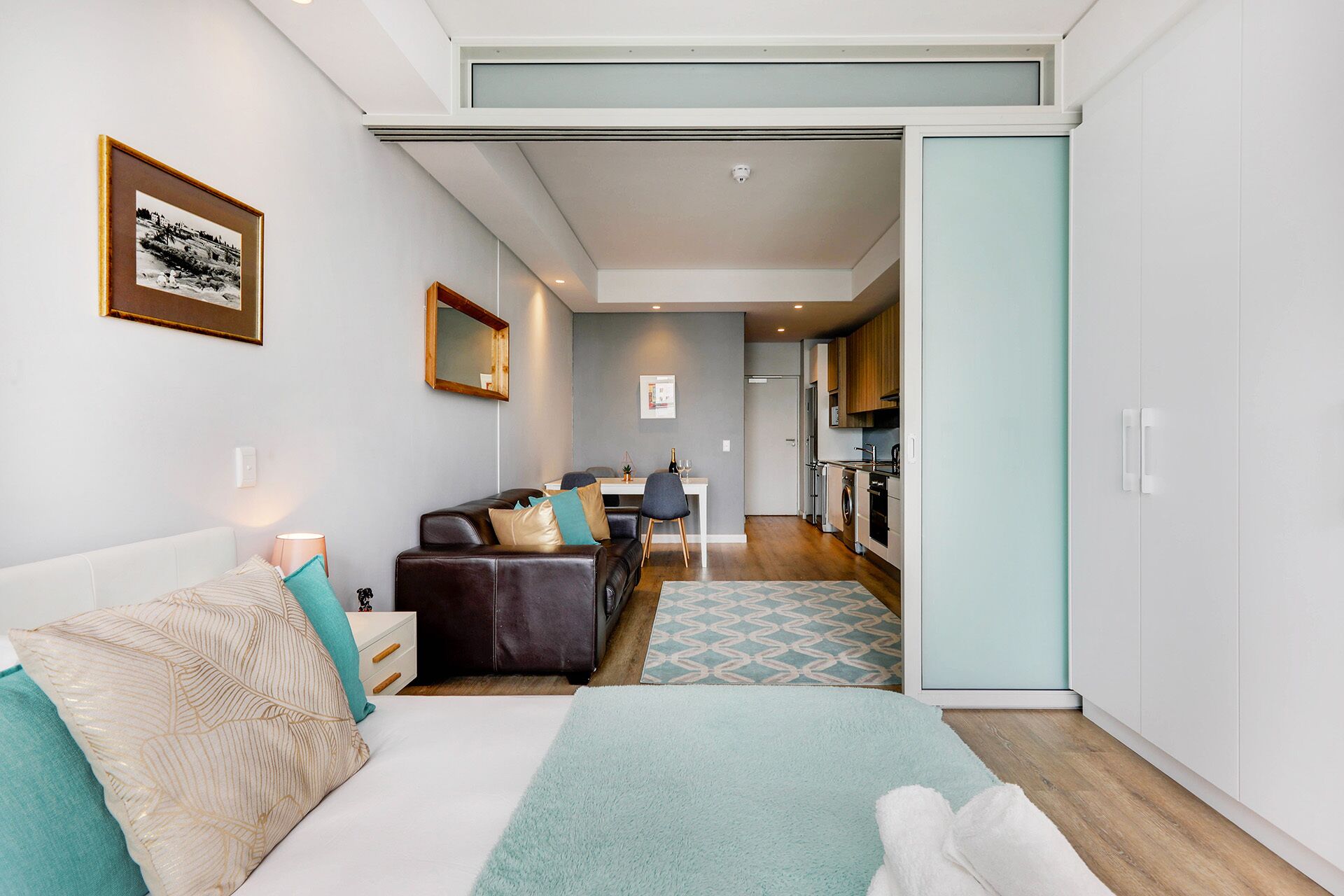 A modern hotel room with a bed, a sofa, a dining area, and a kitchenette.