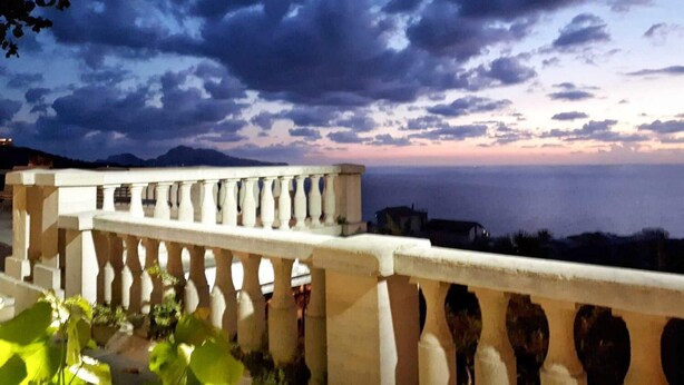 Un balcone con ringhiera che si affaccia su un panorama mozzafiato tra mare e montagne al crepuscolo.