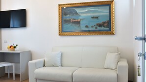 Un soggiorno con un divano bianco, un tavolino con un cesto di frutta e un quadro raffigurante una scena costiera.