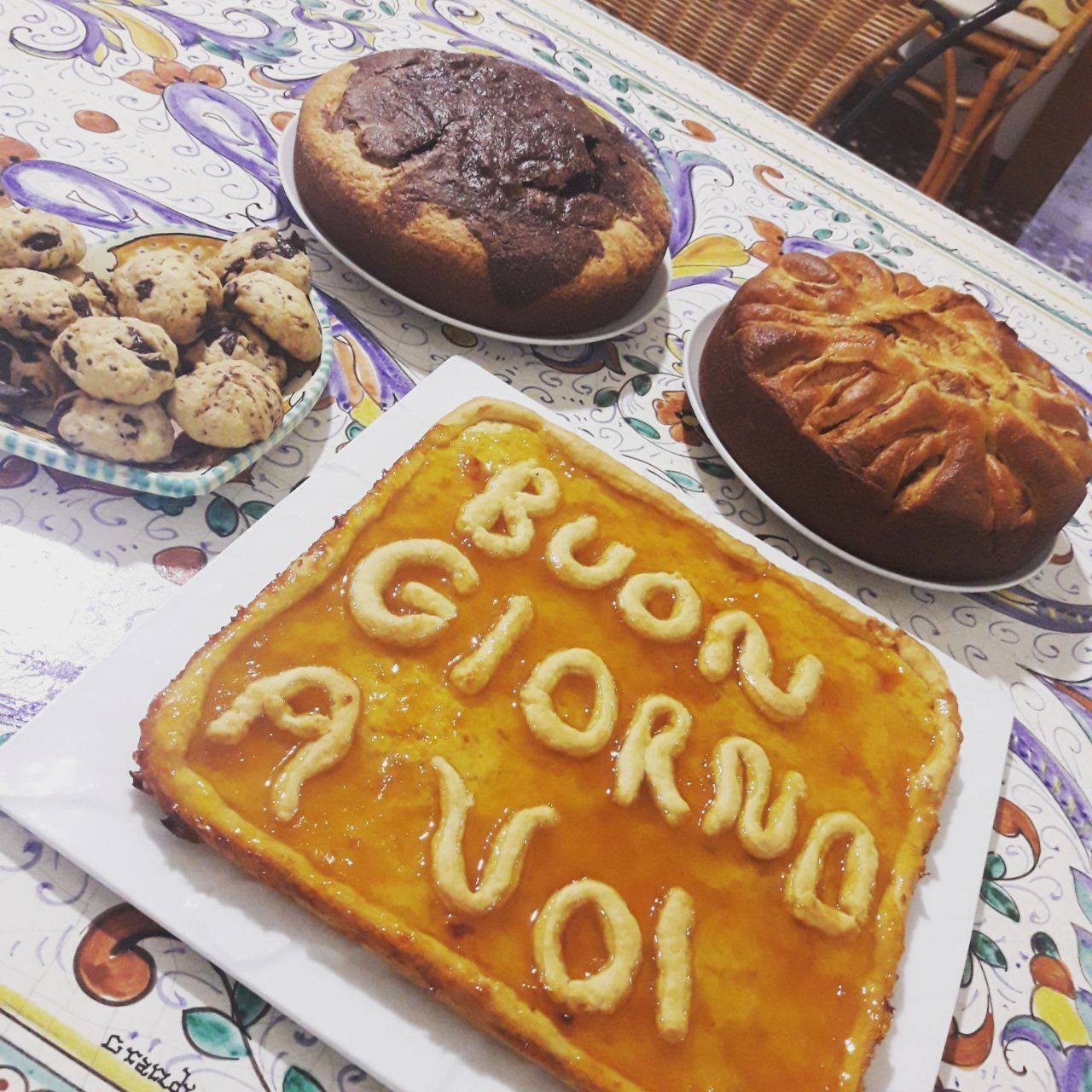 A table with various baked goods, including a rectangular tart with "Buon Giorno AVOI" written on it, a round cake, and a plate of cookies.