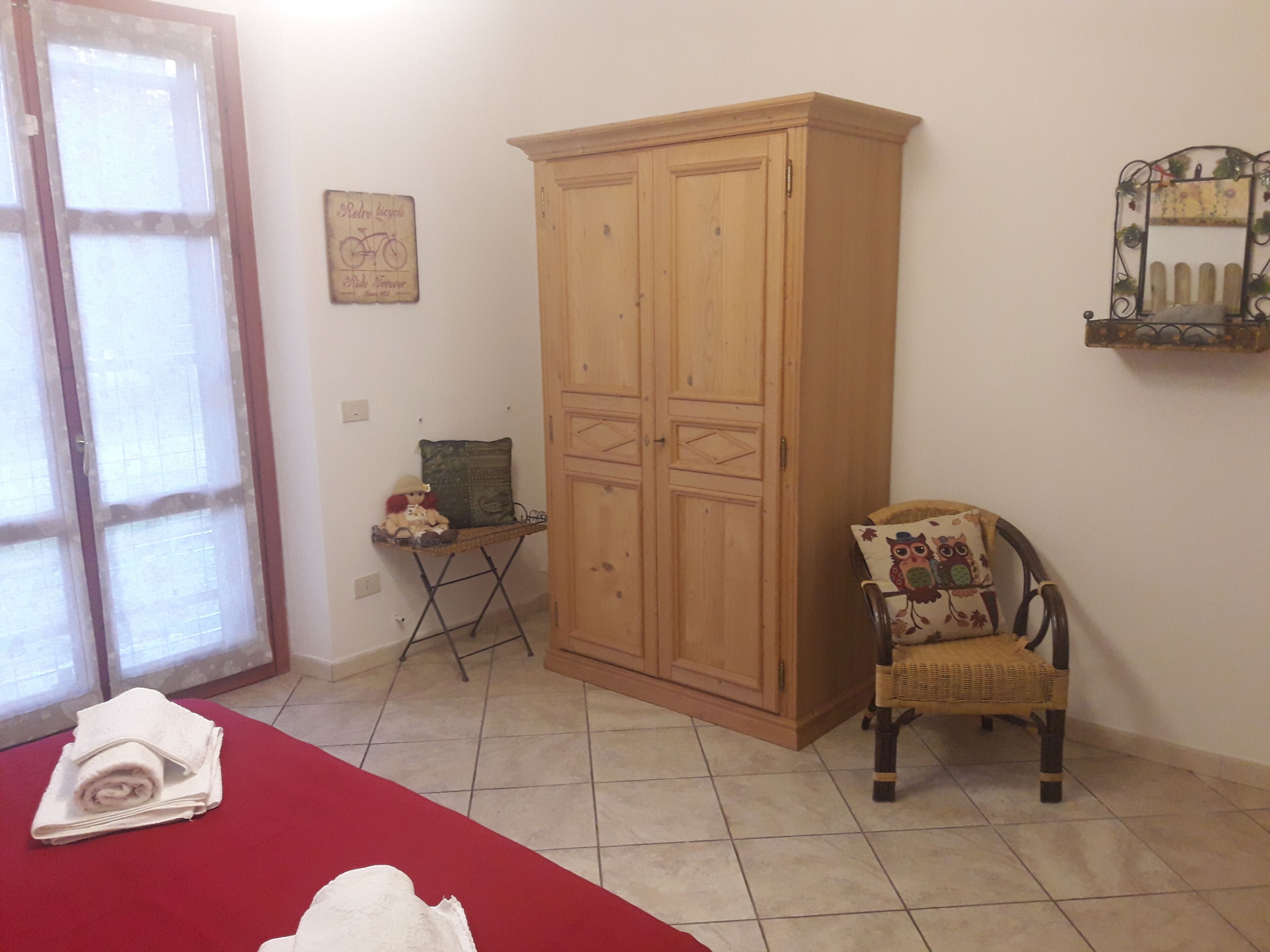 Una habitación con un armario de madera, una silla de mimbre con un cojín con forma de lechuza, una mesita con un cojín, y una colcha roja.