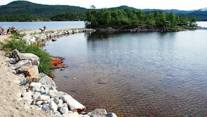 Escena junto al lago con orilla rocosa, agua cristalina y una colina boscosa al fondo.