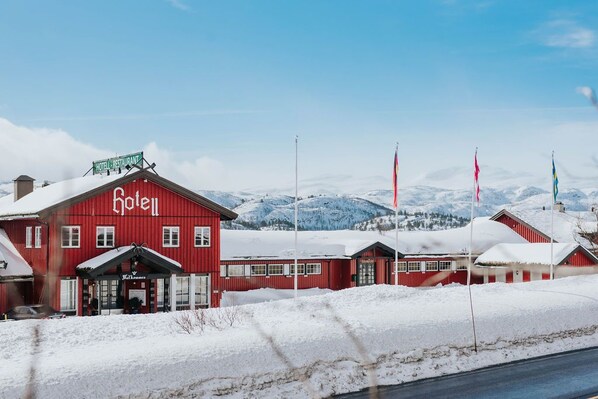 A red hotel building with a sign reading "Hotel Restaurant" and a view of snow-covered mountains in the background. - Vågslidtun Hotell (Vinje)