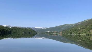 Un lago tranquilo con un reflejo claro, rodeado de colinas boscosas y algunos edificios a lo lejos.
