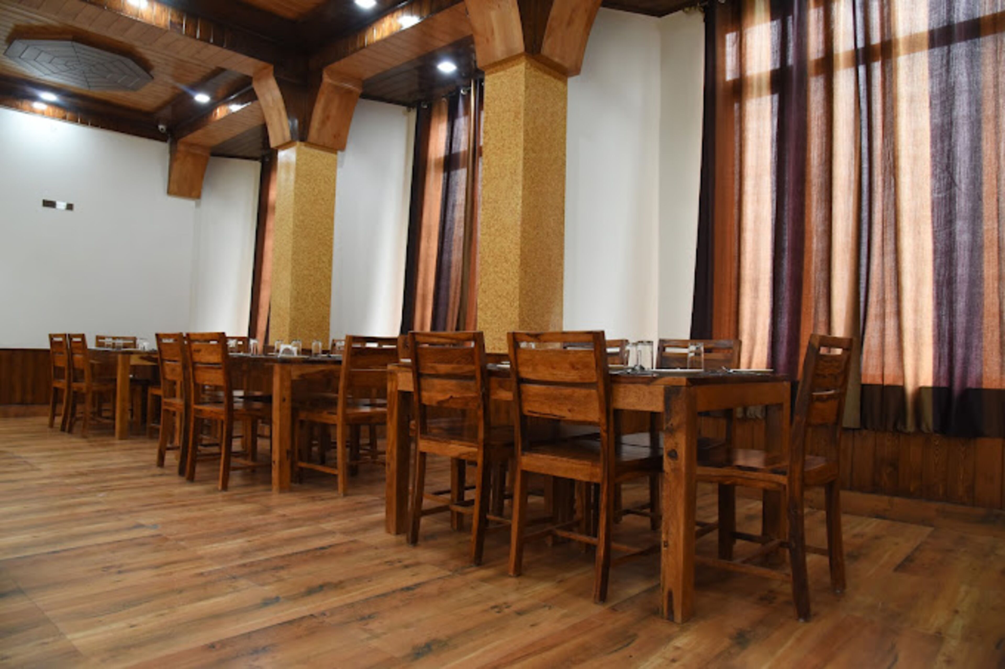 A dining area with wooden tables and chairs, high ceilings with exposed beams, and large windows with curtains.