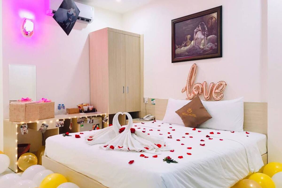 A hotel room with a bed adorned with rose petals and a heart-shaped towel, a minibar, a desk with a photo collage, and a 'Love' balloon decoration.
