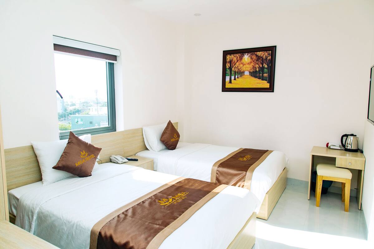 A hotel room with two beds, a desk, a chair, a window with a view of buildings, and a framed picture on the wall.