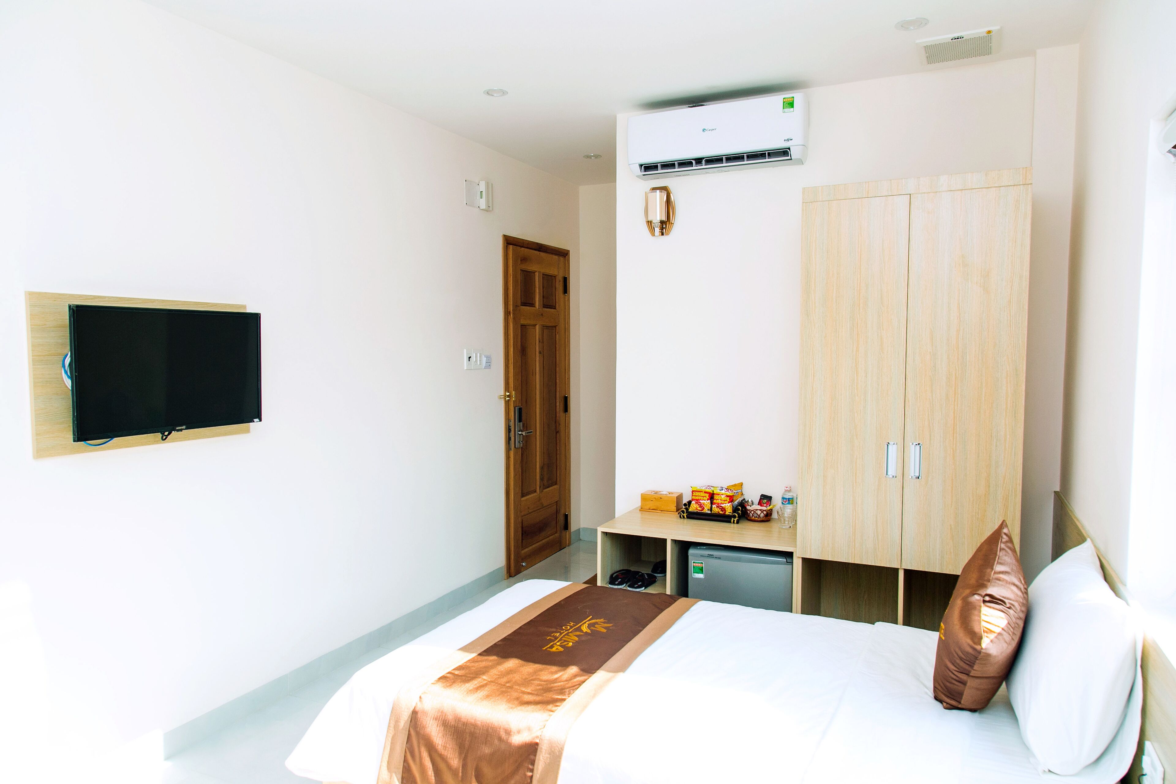 A hotel room with a bed, a television on the wall, a wardrobe, a desk with a chair, and an air conditioning unit.