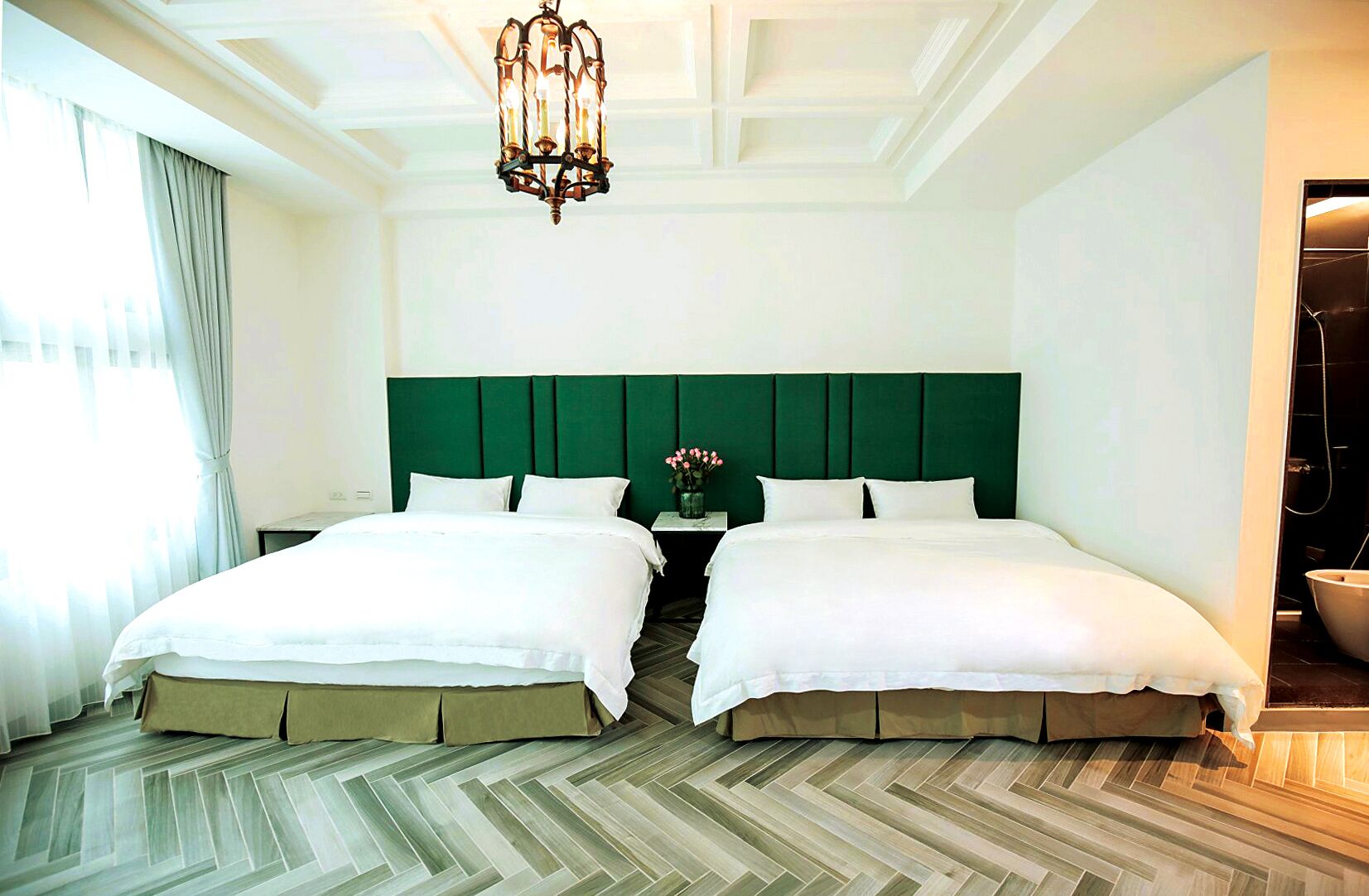 A hotel room with two beds, a green headboard, a wooden herringbone floor, a chandelier, and a window with curtains.