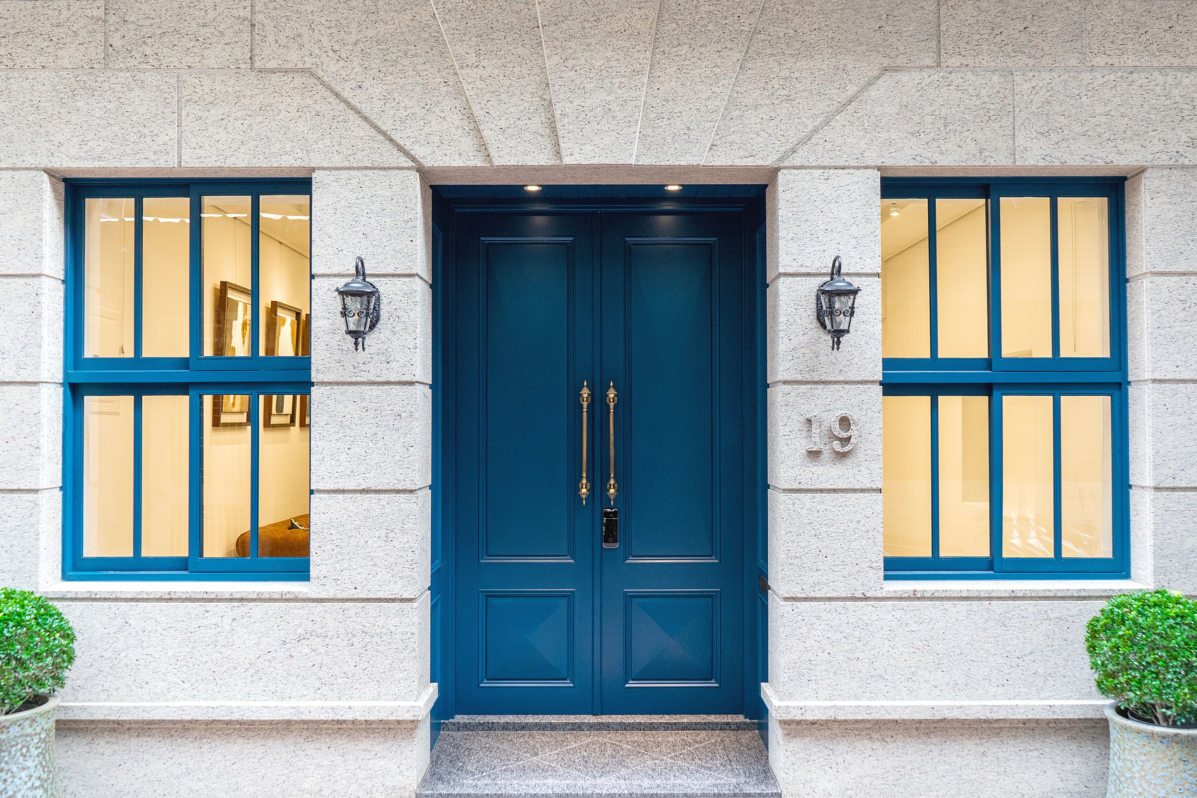 Uma porta dupla azul com o número 19 acima, com duas janelas laterais de moldura azul e interiores iluminados.