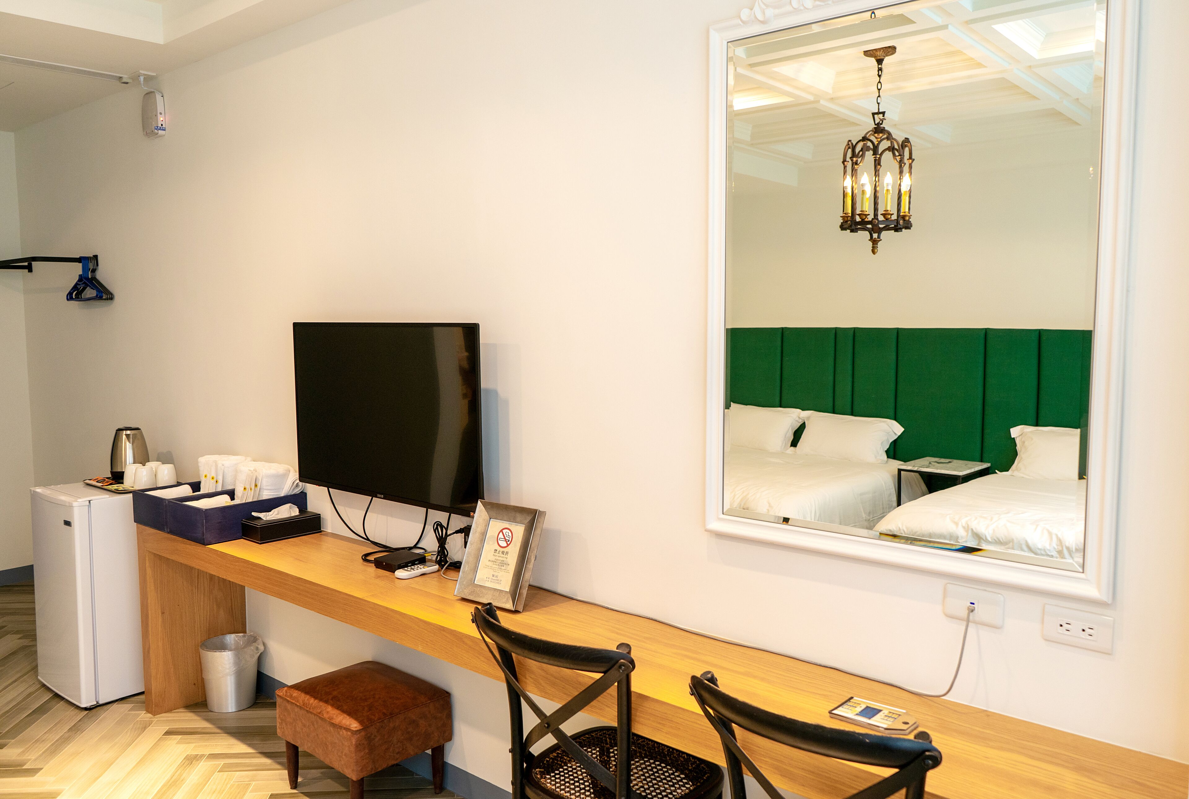 A hotel room with a wooden desk, a television, a small refrigerator, a trash bin, and a large mirror reflecting a green upholstered headboard and a chandelier.
