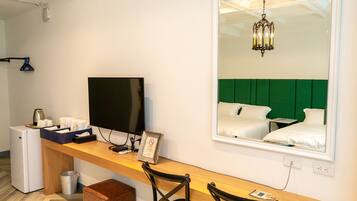 A hotel room with a wooden desk, a television, a small refrigerator, a trash bin, and a large mirror reflecting a green upholstered headboard and a chandelier.