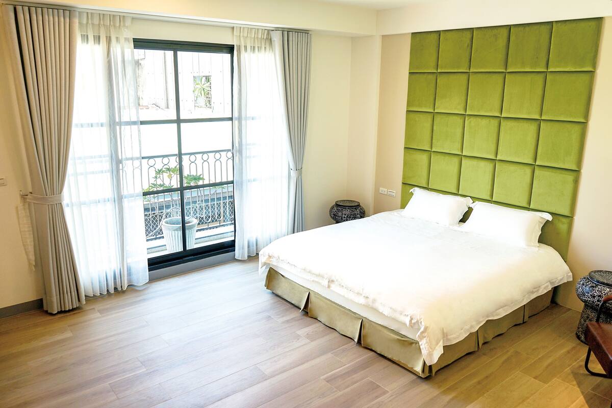 Ein Schlafzimmer mit einem großen Bett, einem gepolsterten, grünen Kopfteil und Blick auf einen Balkon durch ein mit Vorhängen drapiertes Fenster.