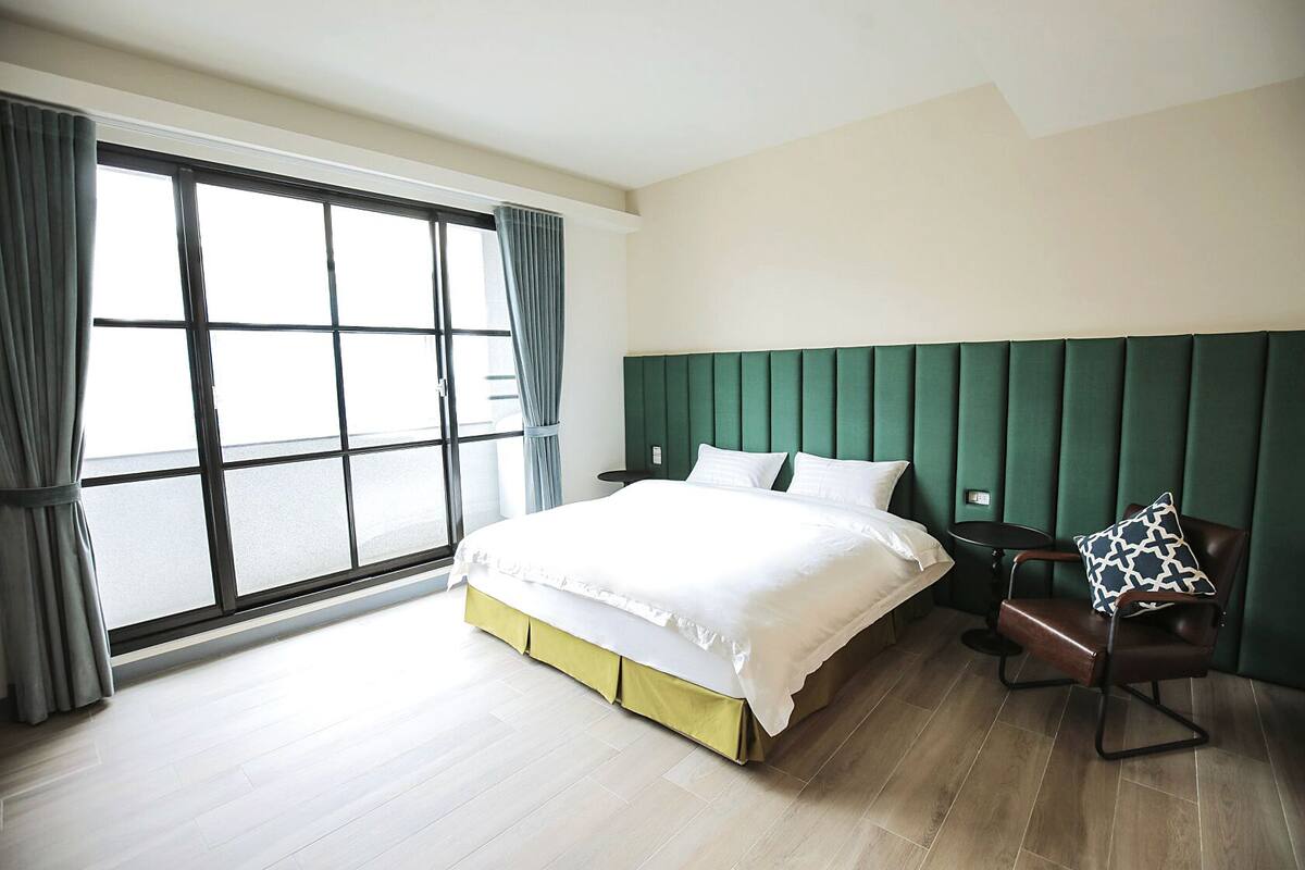 Ein Hotelzimmer mit einem großen Bett, einem grünen Kopfteil, Holzboden und Blick durch ein großes Fenster mit Vorhängen.