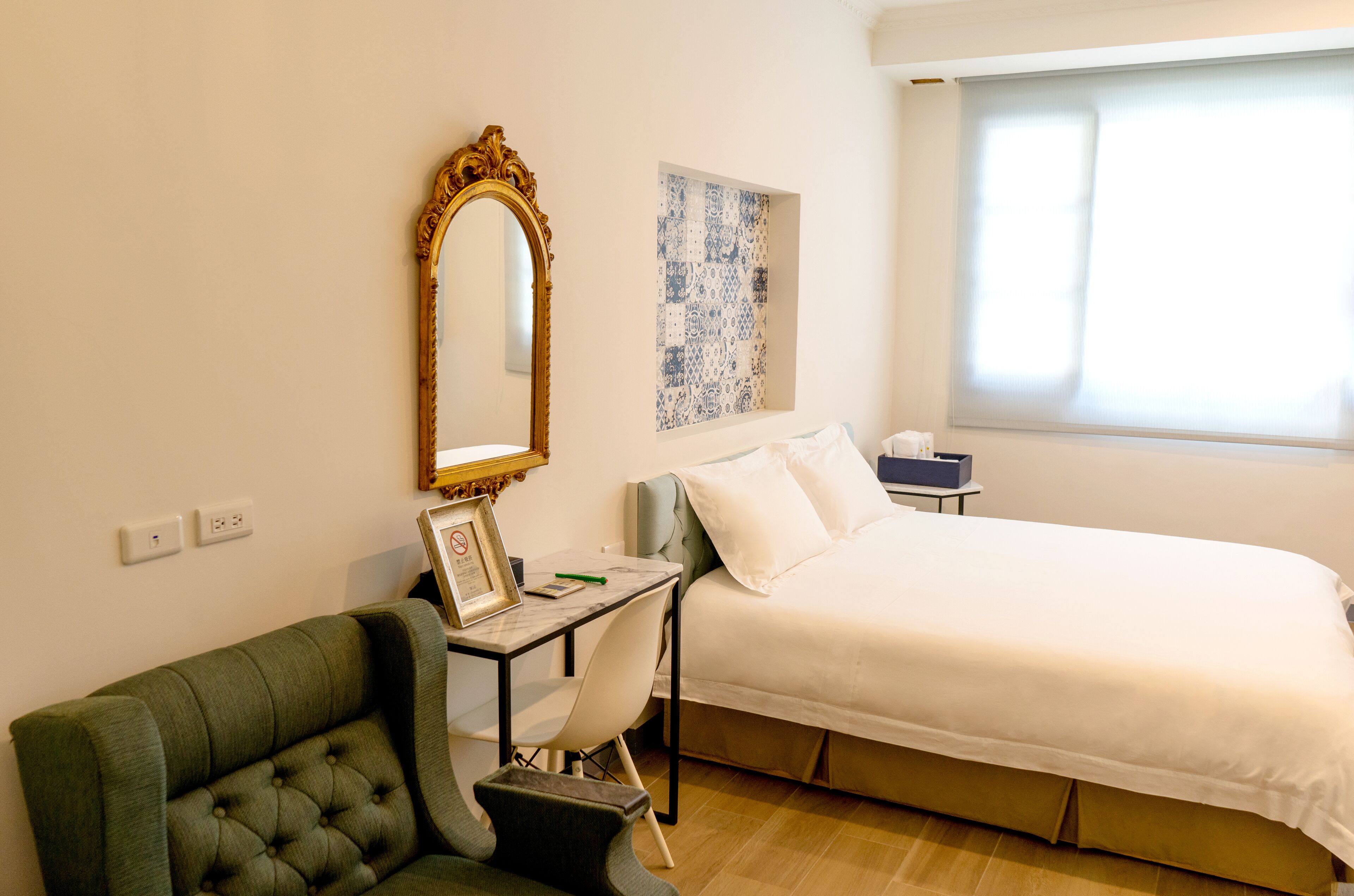 Habitació d'hotel amb llit, escriptori amb cadira, butaca verda, un mirall gran i una finestra amb persianes.