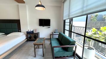 Una habitación de hotel moderna con una cama, un sofá verde, una mesa de madera, un televisor montado en la pared y un amplio ventanal con vistas al exterior.