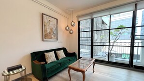 Un salon avec un canapé vert, une table basse en bois et une grande fenêtre donnant sur des immeubles et un balcon.