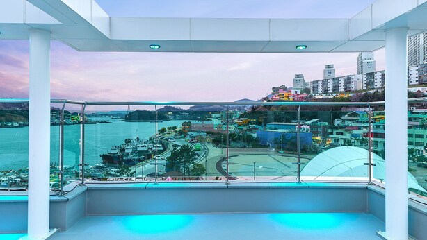A high-rise hotel room with a balcony overlooking a harbor, cityscape, and a sports field. - I Stayed Pension Yeosu (Yeosu)