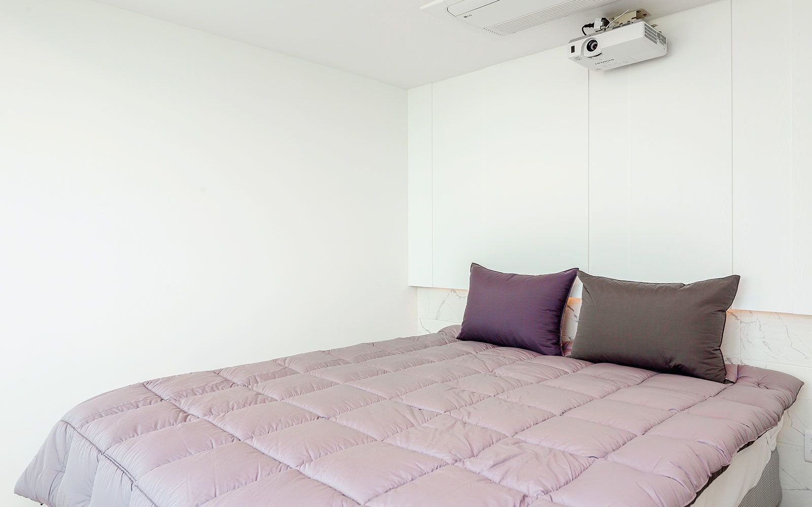 Ein Bett mit gesteppter Bettdecke und zwei Kissen, ein an der Decke befestigter Projektor und eine weiße Wand.