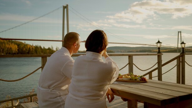 A couple sitting at a wooden table with a view of a bridge and water. - Björkudden Hotell & Restaurang (Sandoverken)