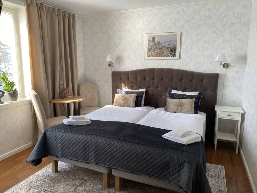 Hotellihuone, jossa on suuri sänky, kaksi yöpöytää, tuoli, ikkuna verhoilla ja seinälle ripustettu taulu.