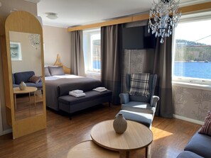 A modern hotel room with a bed, a sofa, a chair, a small table, and a large window offering a view of a body of water. - Björkudden Hotell & Restaurang (Sandoverken)