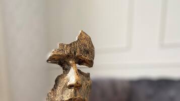 En skulptur på ett gyllene bricka med en svart fot, placerad mot en suddig bakgrund av ett rum med en soffa och ett bord.
